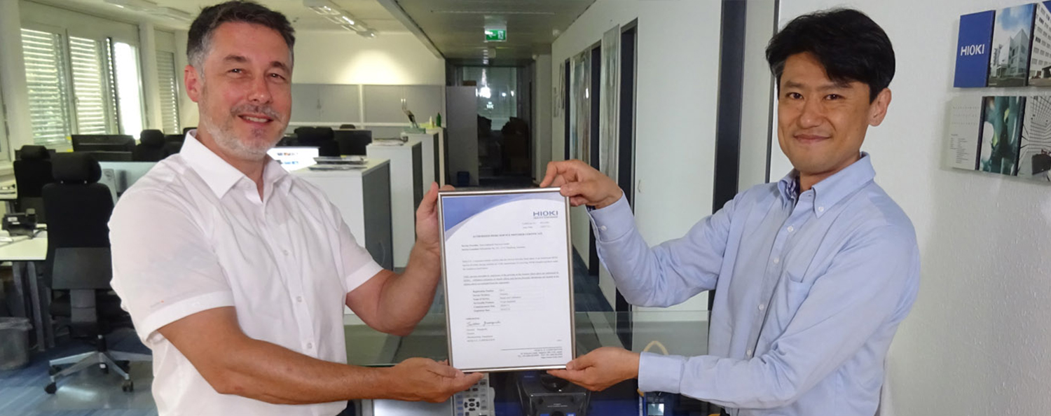 Unsere Zusammenarbeit mit Hioki Zwei Mitarbeiter halten die Urkunde der Zusammenarbeit lächelnd in die Kamera.